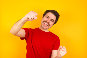 Young Caucasian man wearing red t-shirt standing against yellow background imagine steering wheel helm rudder passing driving exam good mood fast speed