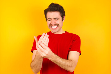 Young Caucasian man wearing red t-shirt standing against yellow wall Suffering pain on hands and fingers, arthritis inflammation