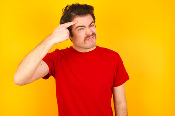 Fototapeta na wymiar Young Caucasian man wearing red t-shirt standing against yellow wall pointing unhappy at pimple on forehead, blackhead infection. Skincare concept.