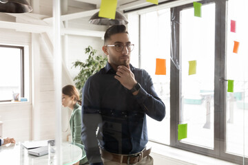 Thoughtful Arabian businessman wearing glasses looking at glass wall with colorful sticky papers, touching chin, pensive executive working on corporate project plan or strategy in creative office