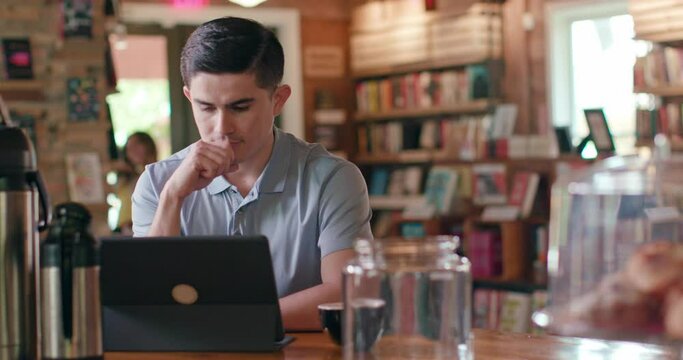 Portrait of young man sitting at table in coffee shop bookstore using a tablet, while a patron walks around the background.