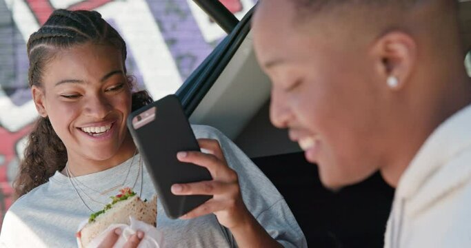 Young African American couple having impromptu picnic lunch in the back of their vehicle, taking selfie with mobile phone 