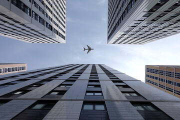 Big airplane flying high above modern city skyscraper buildings with many windows in business cluster view bottom up on bright sunny day