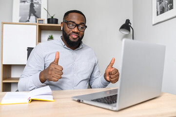 An intelligent young African-American teacher approves the project online on the laptop, wearing glasses, making notes, showing thumbs-up. An excited candidate is on a video call at the job interview