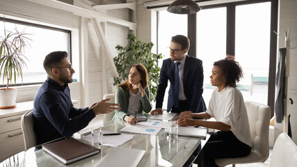 Diverse employees listening to Arabian colleague at meeting in boardroom, businessman wearing glasses sharing ideas with coworkers, business partners discussing project statistics or strategy