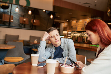 Two teenagers spending time together, sitting in a cafe on a daytime. Displeased guy looking at his girlfriend while she is ignoring him, using her smartphone