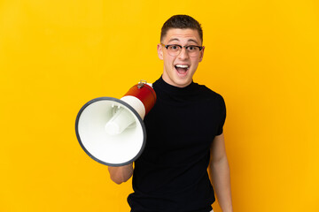 Young caucasian man isolated on yellow background holding a megaphone and with surprise expression