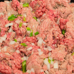 Raw ground meats sitting on a bed of raw onions and green bell peppers in non-stick skillet on a stove top in a domestic kitchen.