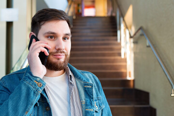 Portrait young man making call on hallway stairs. Caucasian bearded business man in modern city office have mobile conversation, talking by phone indoors.