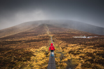 Boy in a red jacket, hiking on wooden path leading through the wicklow mountains, Djouce pek Ireland. Wooden path in foggy mountain landscape, in Autumn. 2019 Ireland