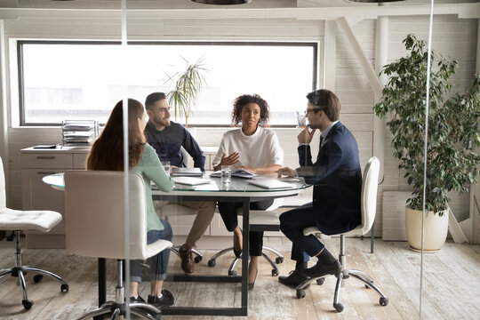 Diverse Business People Partners Discussing Project, Contract Terms, Brainstorming In Modern Board Room Behind Glass Wall, Colleagues Sitting At Table In Office, Talking, Sharing Startup Ideas