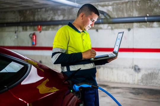 Male technician using mobile phone with laptop by electric car in auto repair shop