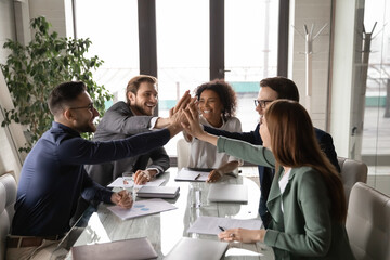Excited diverse business team giving high five at briefing, sitting at table in boardroom, motivated for shared success, overjoyed colleagues joining hands, engaged in team building activity