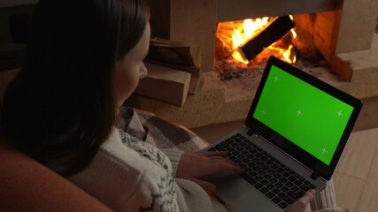 Woman at home, sitting on a sofa by the fireplace, on a laptop celebrates online success with a green layout screen. 