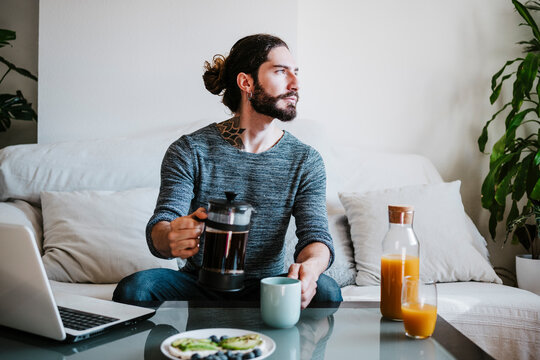 Man with coffee maker looking away while sitting by breakfast on table at home
