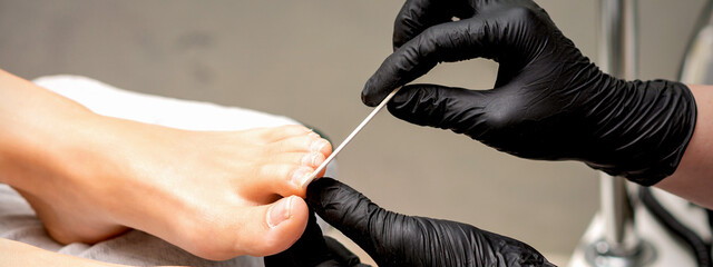 Pedicure master's hands in protective rubber gloves file female toenails with a nail file in a beauty salon © okskukuruza