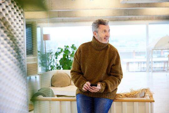 Mature man with mobile phone looking away while sitting on sofa at home
