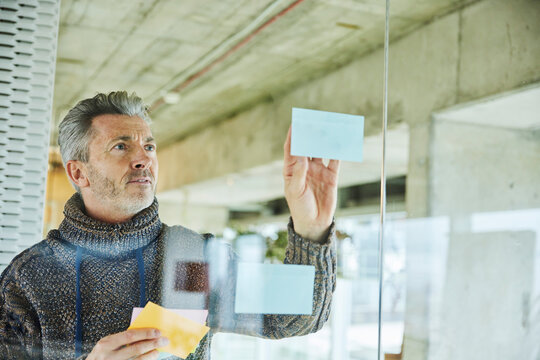 Male businessman putting sticky note while standing by glass wall at office