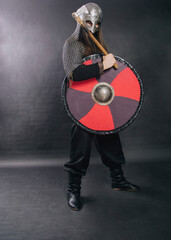 Fototapeta na wymiar Viking stands with an ax on a gray background. A man in chain mail and a helmet holds a weapon. Medieval warrior in armor.