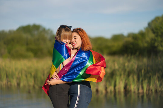 Young lesbian couple covered in rainbow flag embracing each other while standing by lake