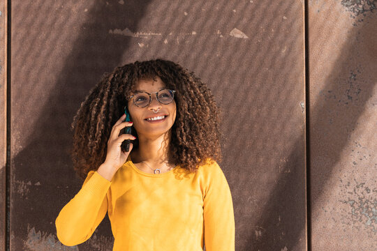 Smiling young woman talking on mobile phone against wall on sunny day