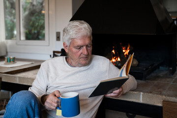 Senior man with coffee cup reading book while sitting by fireplace at home