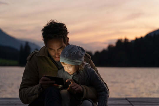 Father and little daughter playing with smart phone on lakeshore jetty at dusk