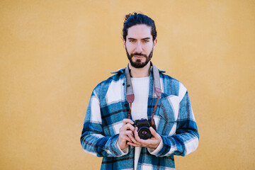 Young man holding camera while standing against yellow wall