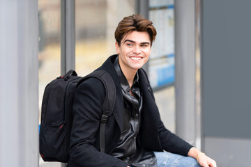 Smiling handsome young man looking away while sitting with backpack at bus stop