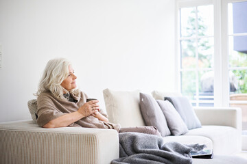 Thoughtful woman with long white hair holding coffee cup while sitting on sofa at apartment