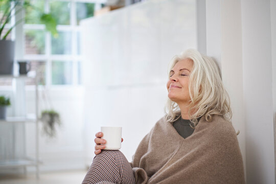 Retired senior woman holding coffee cup while sitting with eyes closed day dreaming at home