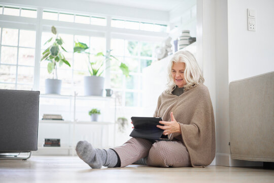 Senior retired woman sitting on floor using digital tablet in living room at apartment