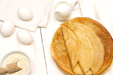 A stack of thin pancakes on a light background with the ingredients for their preparation.