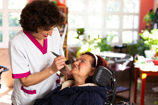 Smiling female nurse doing make-up of disabled woman in nursing home