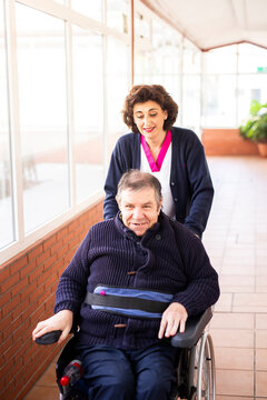 Mature female nurse helping disabled man sitting on wheelchair in corridor of rehabilitation center