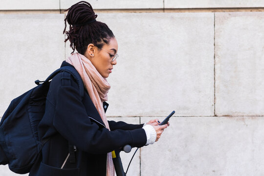 Jamaican woman with dreadlocks using smart phone while leaning on push scooter by wall