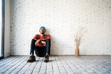 Man wearing knit hat day dreaming while sitting by dried plat vase against white brick wall