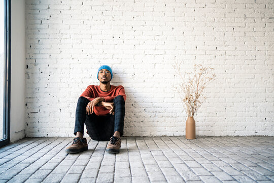 Fototapeta Man wearing knit hat day dreaming while sitting by dried plat vase against white brick wall