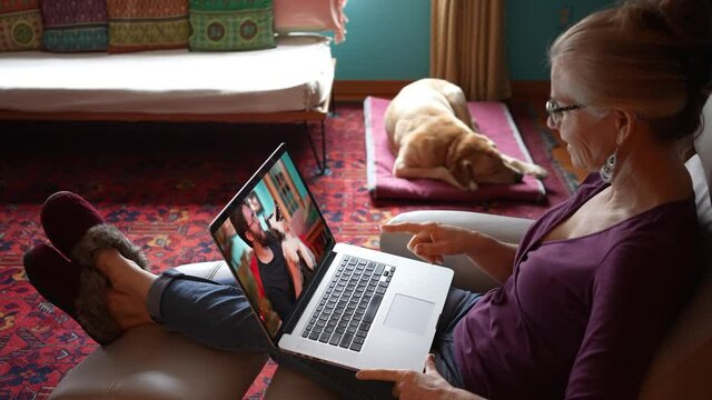 Woman smiling with her feet with slippers up while having video chat with man with Husky dog on her laptop computer in living room. Concept of remote social distancing.