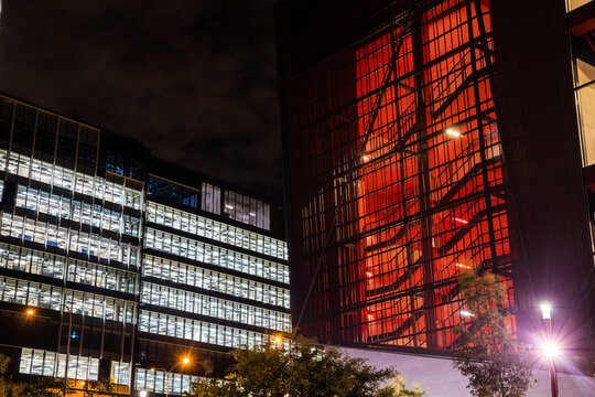 Office Building At Night, Windows Illuminated White And Stairwell Of Other Building Illuminated Red