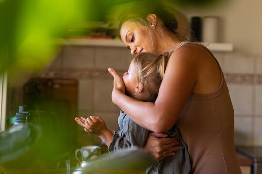 Young Mum Wiping Child's Face At Kitchen Sink Glimpsed Through Blurred Greenery