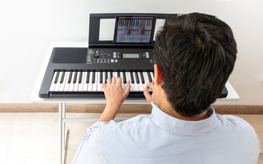 Back shot of a man in a light blue shirt playing the piano using an application on a tablet to learn music.