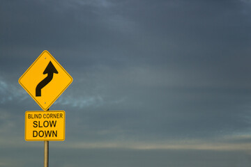 Blind corner slow down road sign against dark clouds