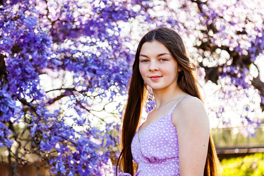Happy brunette teenage girl standing among flowering branches of purple jacaranda tree