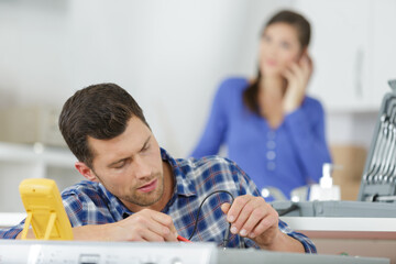 serviceman testing appliance with multimeter housewife in background