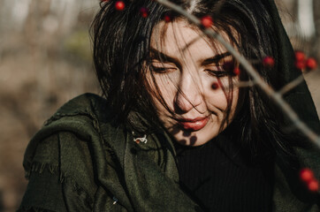 Coquette close up portrait of a young woman looking down, wearing black and a dark green scarf over shoulders, winter berries branch on foreground