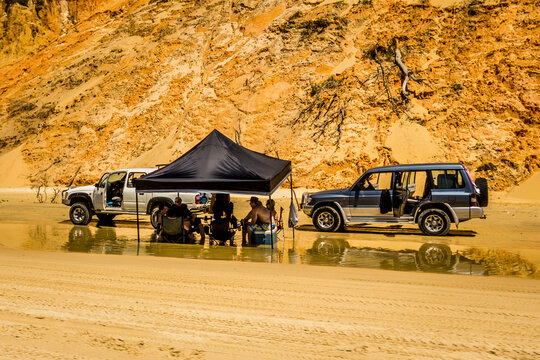 Group of people sit in shallow water beside four wheel drive vehicles