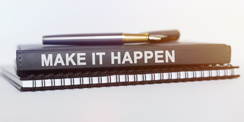 On the table are a notebook, a pen and a book. The book says - MAKE IT HAPPEN