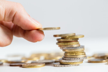 Holding coin in fingers over the stack of money from close up. Adding one euro on pile of metalic...