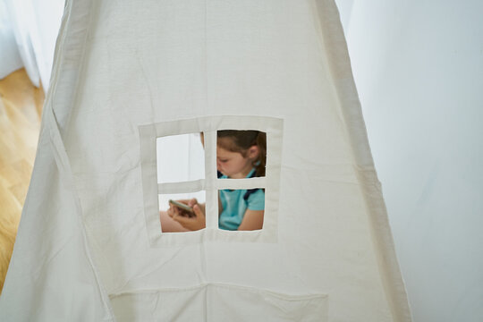 Little girl looking at her smartphone inside a white teepee tent inside her house. Technology concept